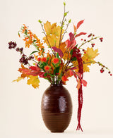 I en brun vase er 'Rustic Glow'-buketten af ​​kunstige silkeblomster, med gule, orange og røde blade, røde bær, orange blomster og lange, hængende røde amaranthus mod neutral baggrund.