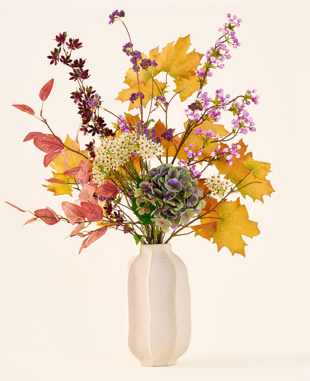 'Wild Ametyst'-buketten af ​​kunstige silkeblomster, med efterårsblade, lilla og hvide blomster og grønne hortensiaer, er i en hvid keramikvase mod en almindelig, lys baggrund.