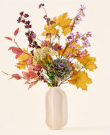 'Wild Ametyst'-buketten af ​​kunstige silkeblomster, med efterårsblade, lilla og hvide blomster og grønne hortensiaer, er i en hvid keramikvase mod en almindelig, lys baggrund.