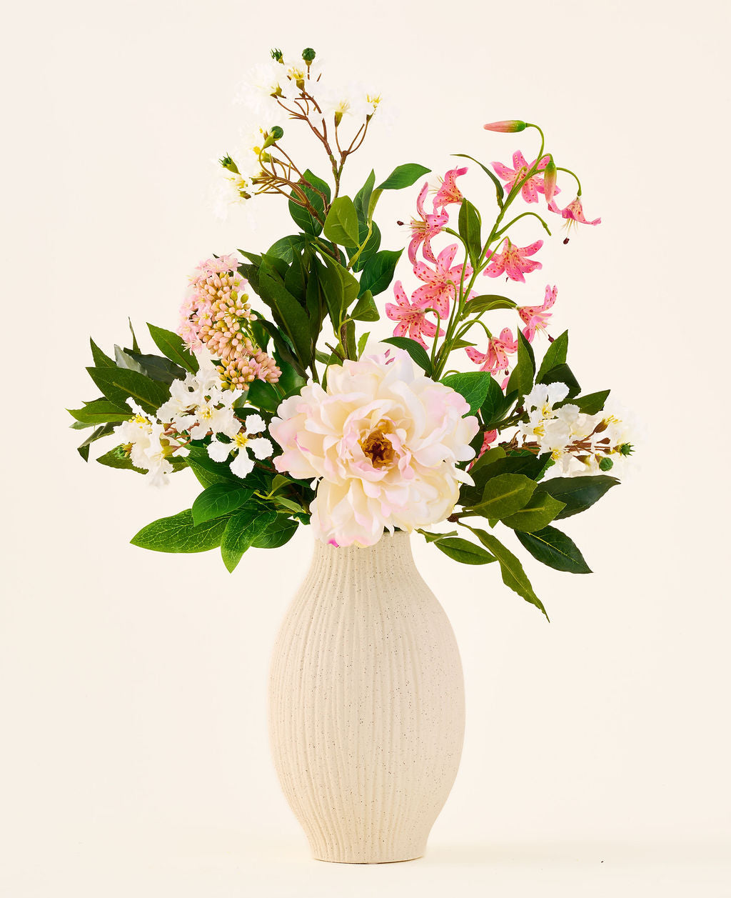 I en cremefarvet keramikvase er 'Light & Lovely' buketten med kunstige silkeblomster fra Easyplants, bestående af et blandet arrangement af hvide, lyserøde og lyslilla silkeblomster med grønne blade, mod en almindelig, lys baggrund.