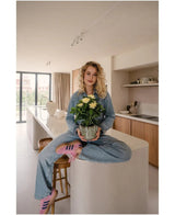 En kvinde med krøllet blond hår, iført denimtøj og lyserøde sneakers, sidder overskrævs på en køkkenø i et moderne, solbelyst køkken og holder den Artificial Gardenia UV 39 cm-plante i hænderne.