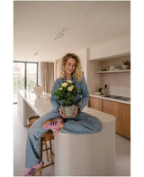 En kvinde med krøllet blond hår, iklædt jeans og stribede sokker, sidder smilende på køkkenbordet, mens hun holder den Artificial Gardenia UV 39 cm plante – en potteplante med gule blomster. I baggrunden ses et moderne køkken med træskabe og hylder.