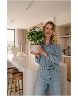 En smilende kvinde i denimoutfit holder den Artificial Gardenia UV 39 cm hvid kunstig plante i et moderne køkken med træskamler, hvide bordplader og neutral indretning.