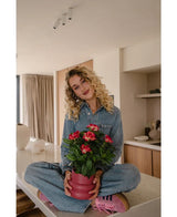 En kvinde med krøllet blond hår, iført en denim jumpsuit og lyserøde sneakers, sidder overskrævs på en køkkenbord og smiler, mens hun holder den Artificial Gardenia UV 39 cm plante i fuchsia.