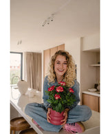 En smilende kvinde med krøllet blond hår sidder overskrævs på en lys, moderne bordplade og holder den Artificial Gardenia UV 39 cm fuchsia-plante – en lyserød potteplante med levende røde blomster. Hun er iført jeans, blå skjorte og lyserøde sko.