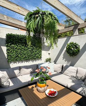En solrig terrasse med beige siddemøbler med hynder og et træbord, dekoreret med en ananasvase med lyserøde blomster og en skål med jordbær. Grønne vægplanter giver et frodigt interiør, suppleret med den kunstige hængeplantebregne UV 60 cm i sort ring som en slående hængende accent.