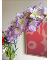 Nærbillede af den kunstige blomst Delphinium 105 cm lilla, arrangeret i en vase, med et lyserødt og rødt blomsterkunstværk på væggen i baggrunden.