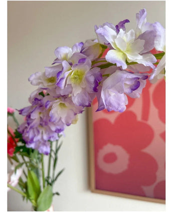 Nærbillede af den kunstige blomst Delphinium 105 cm lilla, arrangeret i en vase, med et lyserødt og rødt blomsterkunstværk på væggen i baggrunden.