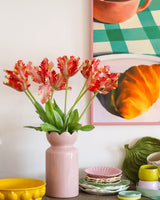 På et bord med farverigt service står en lyserød vase indeholdende den Kunstige Blomst Fransk Parakit Tulipan 76 cm rød/gul, mens der i baggrunden delvist ses et indrammet maleri af en croissant og en kop.
