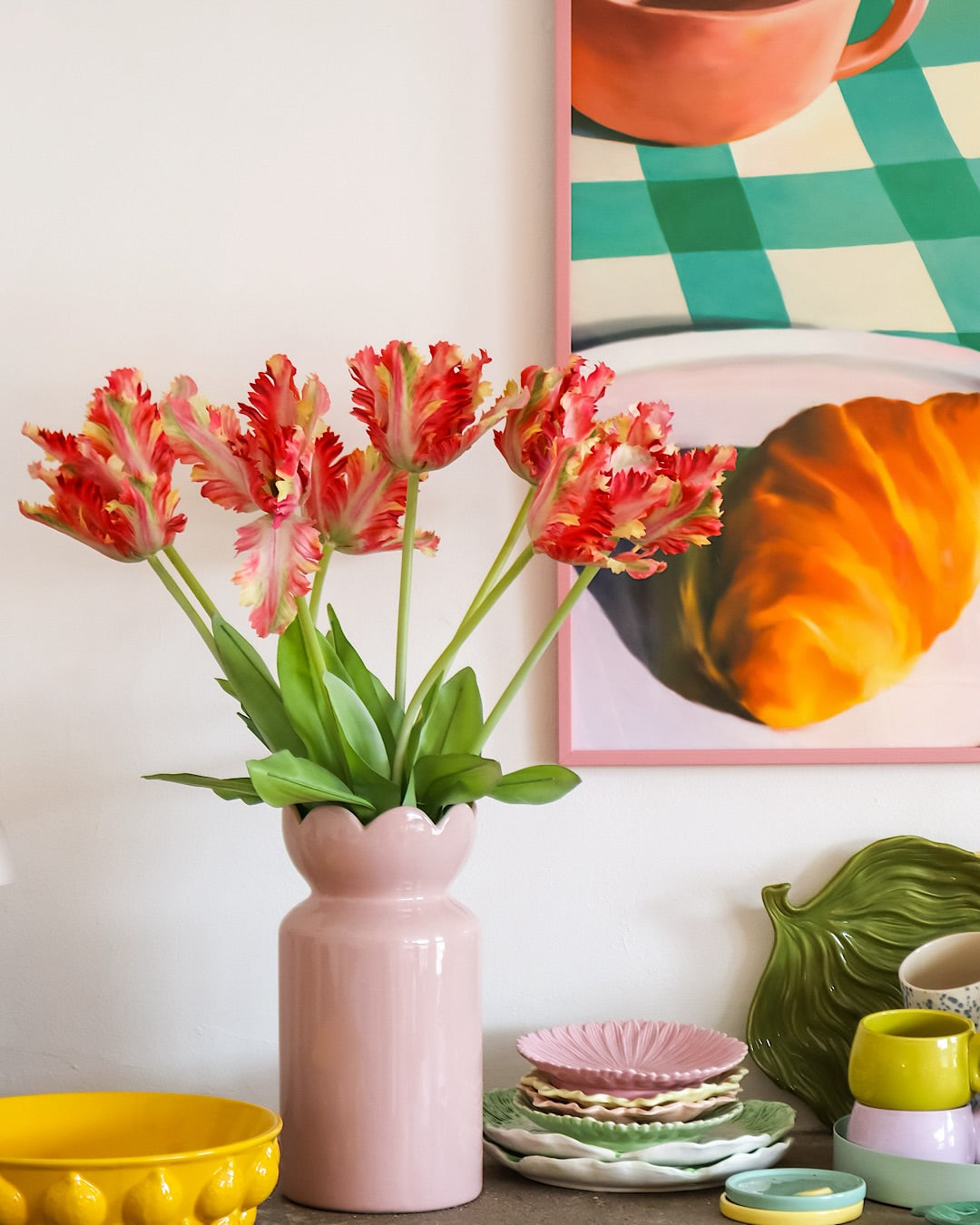På et bord med farverigt service står en lyserød vase indeholdende den Kunstige Blomst Fransk Parakit Tulipan 76 cm rød/gul, mens der i baggrunden delvist ses et indrammet maleri af en croissant og en kop.