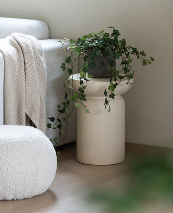 Een Kunst Klimop Deluxe Groen 90 cm staat in een beige vaas op een houten vloer naast een lichtgrijze bank waarop een crèmekleurige plaid ligt en een witte poef met structuur.
