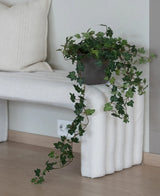 De Kunst Klimop Deluxe Groen 90 cm, met zijn weelderige hangende ranken, staat op een wit bankje met structuur naast een crèmekleurig kussen in een minimalistische kamer met lichte houten vloeren.
