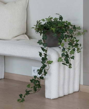 De Kunst Klimop Deluxe Groen 90 cm, met zijn weelderige hangende ranken, staat op een wit bankje met structuur naast een crèmekleurig kussen in een minimalistische kamer met lichte houten vloeren.
