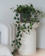 Een Kunst Klimop Deluxe Groen, een hangende klimop van 90 cm in een grijze pot, staat op een modern, wit, sculpturaal voetstuk naast een opgevouwen witte deken en een wit kussen met structuur.