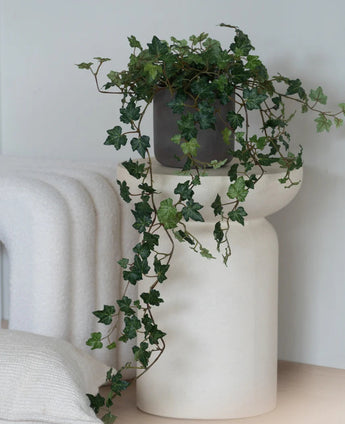 Een Kunst Klimop Deluxe Groen, een hangende klimop van 90 cm in een grijze pot, staat op een modern, wit, sculpturaal voetstuk naast een opgevouwen witte deken en een wit kussen met structuur.