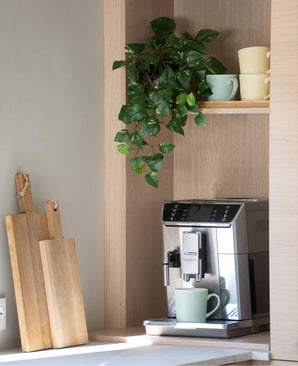 Op het aanrecht staat een zilveren koffiezetapparaat met een groene mok. Daarboven, op een houten plank, staan pastelkleurige kopjes opgestapeld en hangt een ‘Kunst Hangende goudrank 60 cm UV’. Vlak daarnaast staan houten snijplanken tegen de muur geleund.