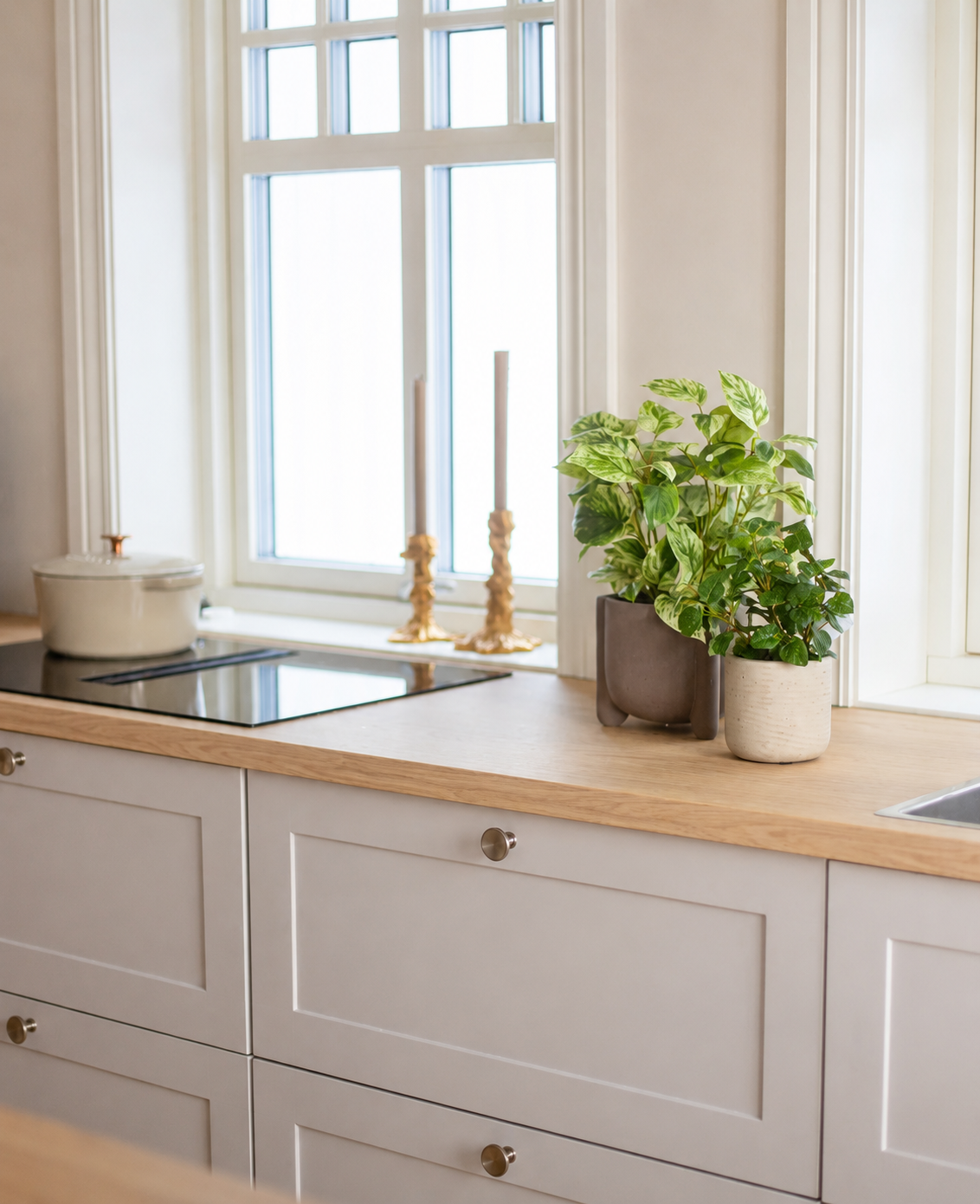 Een lichte keuken met houten werkbladen, witte kasten, twee gouden kandelaars, een witte pan op het fornuis en een groot raam dat natuurlijk licht binnenlaat. De ruimte wordt geaccentueerd door de Kunst Goudrank groen-wit in pot UV 35 cm.