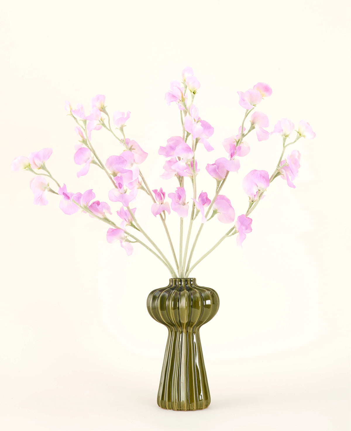 En grøn glasvase viser Silk kunstig blomsterbuket Ært pink med blødt grønt løv, med flere lange stilke og mod en almindelig råhvid baggrund.