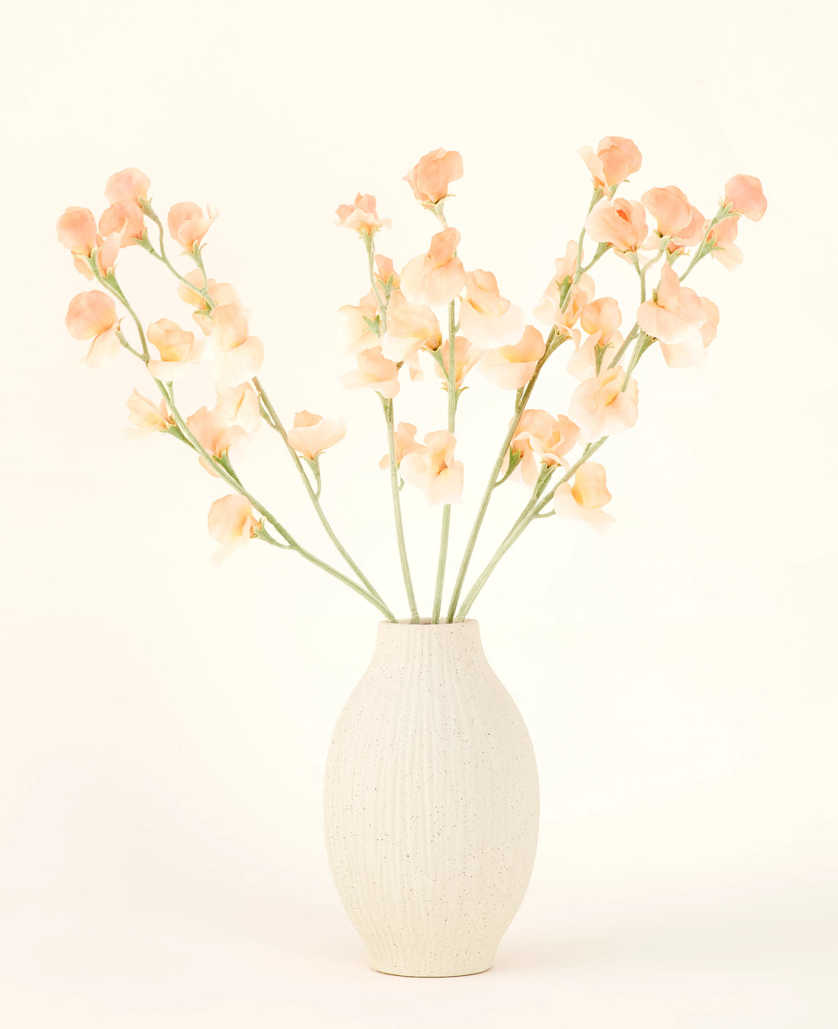 Den cremefarvede keramikvase indeholder Silk kunstig blomsterbuket Pea Peach, med sart fersken og lyserøde blomster med lange grønne stilke mod en lys, neutral baggrund.