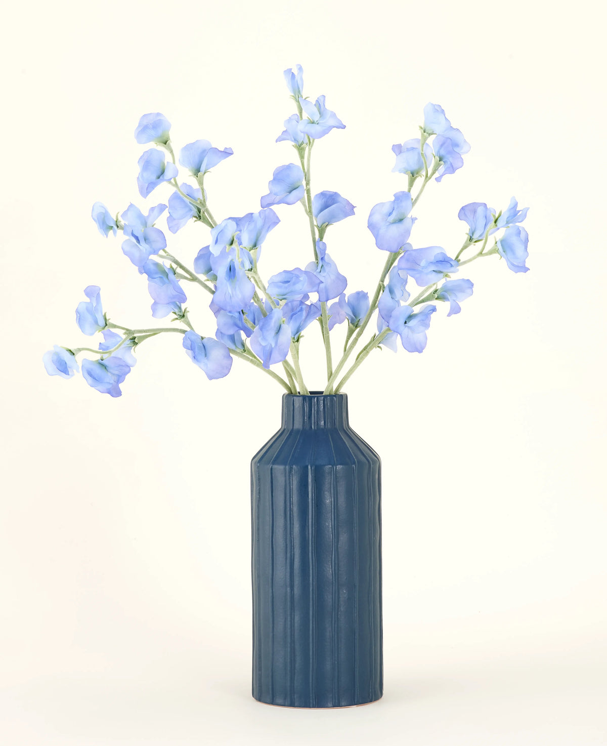 En blå ribbet keramikvase viser Pea Blue kunstige blomsterbuket i silke med sine sarte lyseblå kunstige blomster mod en almindelig, lys baggrund.