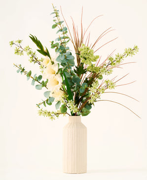In een beige vaas staat het boeket „Wild Ivory“ van Zijden kunstbloemen, met groene bladeren, eucalyptus, witte bloemen, kleine groene bloesems en lange, dunne bruine grassen tegen een lichte achtergrond.