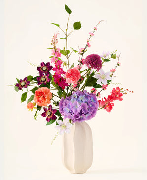 In een witte keramische vaas staat het boeket ‘Rosy Revery’ van Zijden kunstbloemen, een levendig arrangement van roze, paarse, oranje en rode bloemen met groene bladeren en stelen, tegen een lichte achtergrond.