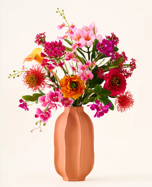 In een perzikkleurige vaas staat het boeket „Tropical Sunset“ van Zijden kunstbloemen, bestaande uit een levendige mix van roze, oranje, gele en rode bloemen tegen een effen, lichte achtergrond.
