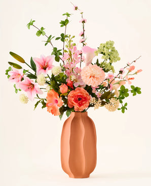In een terracotta vaas staat het kleurrijke boeket ‘Yasmine’s Choice’ van zijden kunstbloemen, bestaande uit roze lelies, perzikkleurige rozen en groen blad, vervaardigd uit hoogwaardige zijden bloemen van Easyplants, allemaal prachtig gearrangeerd tegen een lichte achtergrond.
