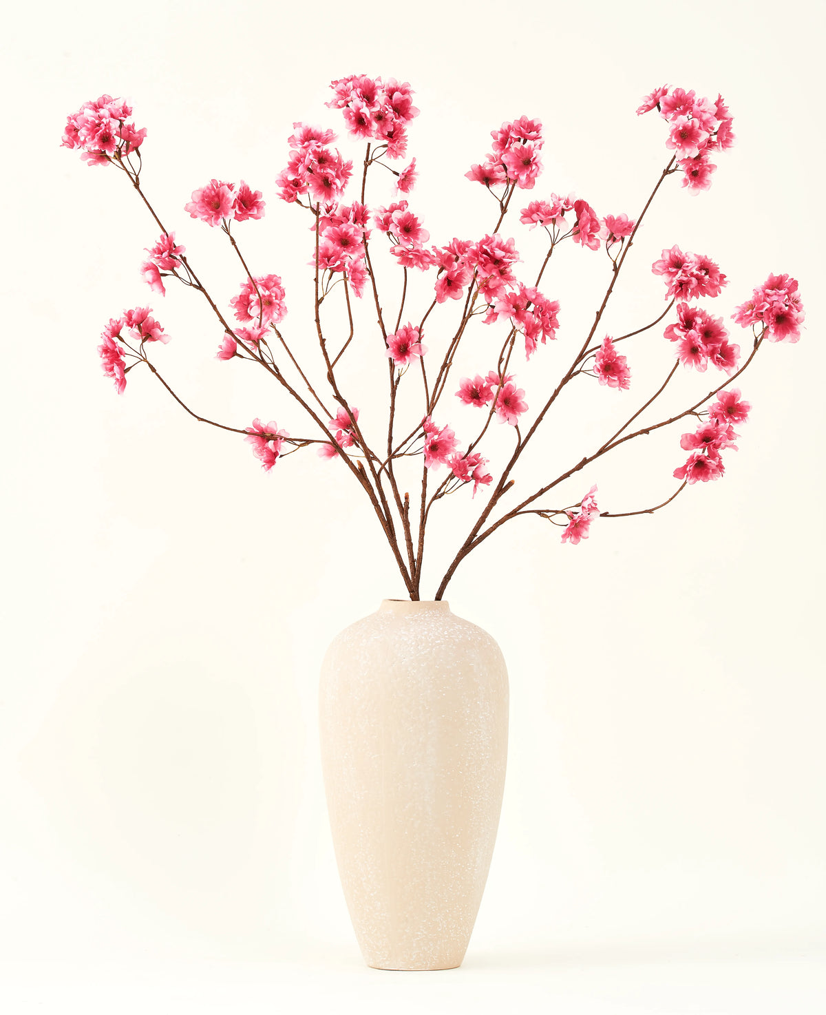 Silke kunstige blomsterbuketten Cherry blossom fuchsia 114 cm tilføjer en levende pop af farve til dit interiør med sine slående grene og fuchsia blomster, smukt udstillet i en beige keramisk vase mod en almindelig råhvid baggrund.