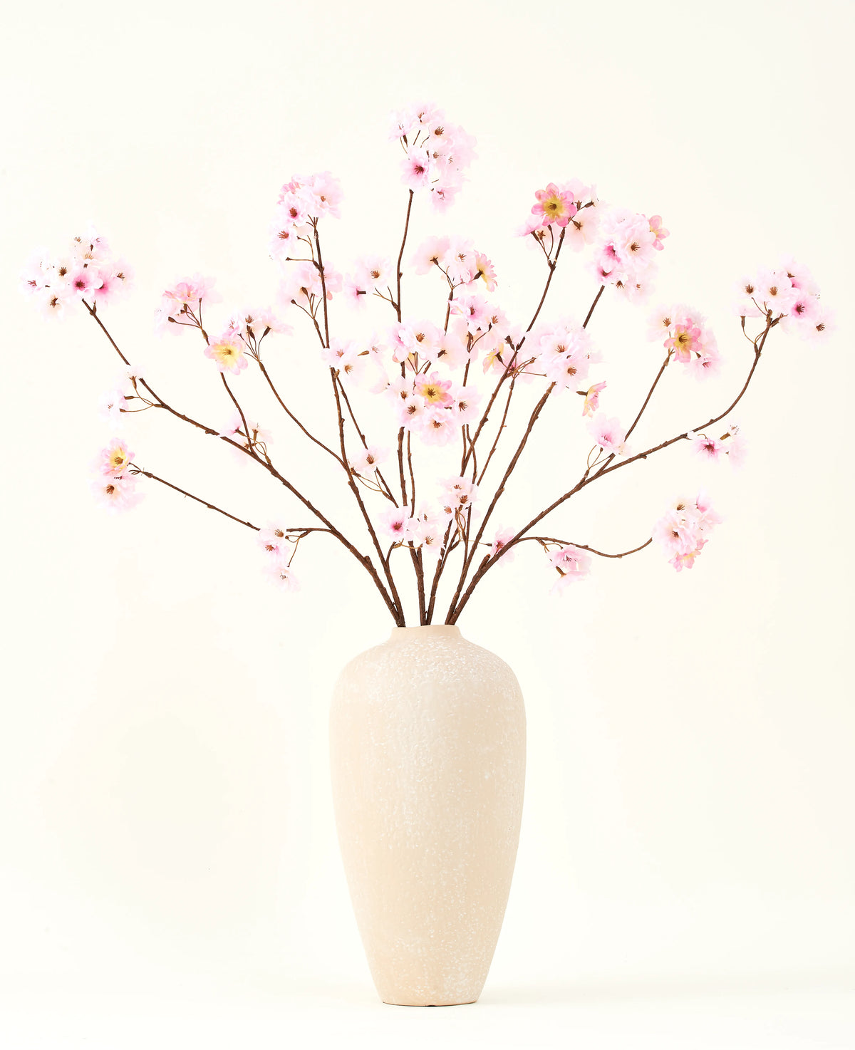 Silke kunstige blomsterbuketten Cherry blossom pink 114 cm har lange, tynde grene med bundter af lyserøde blomster i en beige keramikvase, som danner en slående kontrast mod den almindelige hvide baggrund.