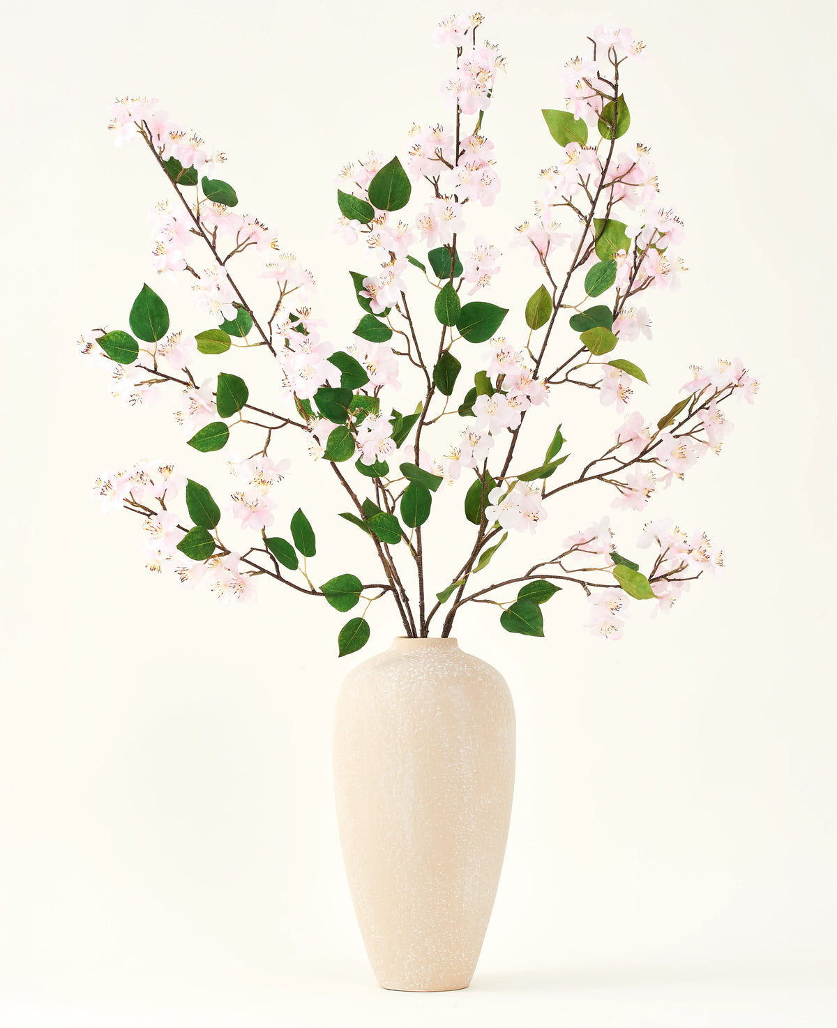 En beige keramikvase indeholder flere lange grene med Silk kunstige blomsterbuket af Peach Blossom pink og grønne blade, mod en almindelig hvid baggrund - perfekt til et kunstigt blomsterinteriør.