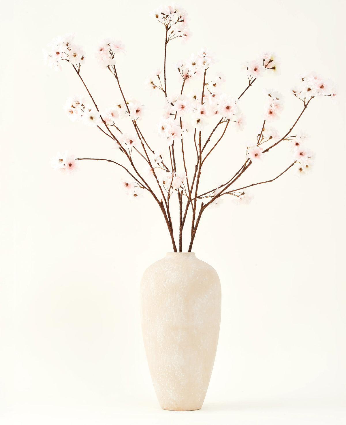 Silke kunstige blomsterbuketten Cherry blossom blush 114 cm har høje, sarte grene i en enkel beige vase, som tilføjer et blødt, elegant touch til dit interiør mod en almindelig, lys baggrund.