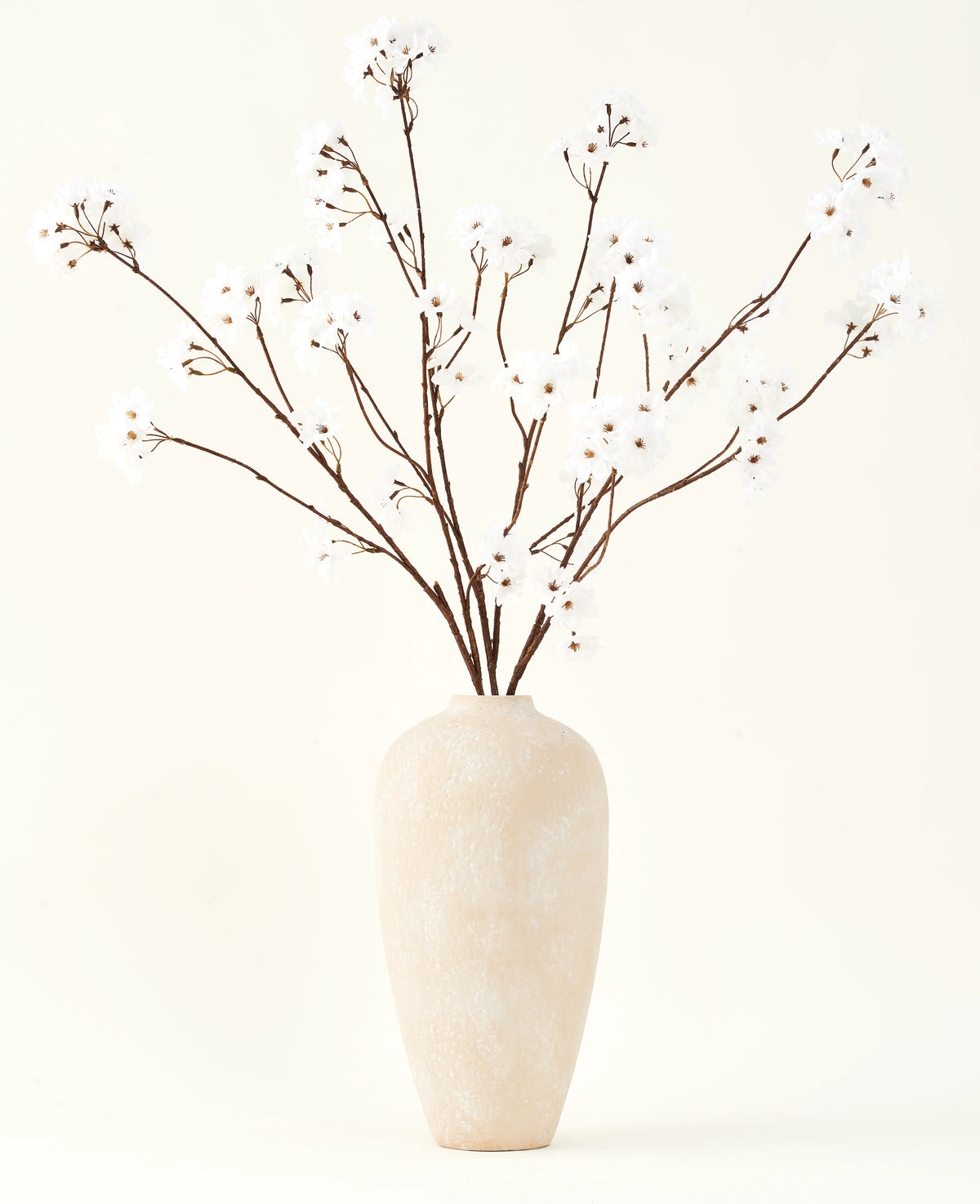 Silke kunstige blomsterbuketten Kirsebærblomstcreme 114 cm står højt i en beige keramikvase og bringer en fredfyldt dekoration til ethvert rum med sine sarte, cremefarvede grene mod en lys baggrund.