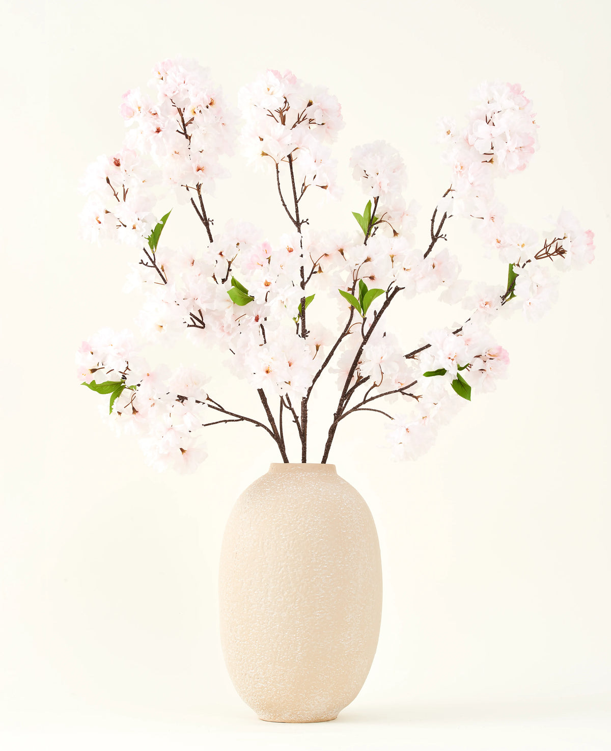 Silke kunstige blomsterbuketten Cherry blossom blush 119 cm viser sarte, lyserøde kirsebærblomster med grønne blade i en beige vase, ideel som en kunstig blomsterdekoration på en lys baggrund.