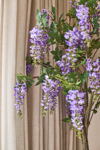 Foran de beige gardiner hænger en kunstig Wisteria-plante på 150 cm med grønne blade.