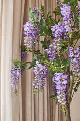 Foran de beige gardiner hænger en kunstig Wisteria-plante på 150 cm med grønne blade.
