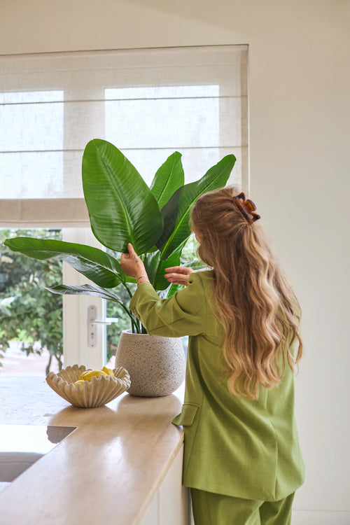 En kvinde med langt bølget hår, klædt i et grønt jakkesæt, plejer en 90 cm Strelitzia Real Touch Deluxe kunstig plante på en disk ved siden af ​​en skål med citroner i et lyst rum med et vindue.