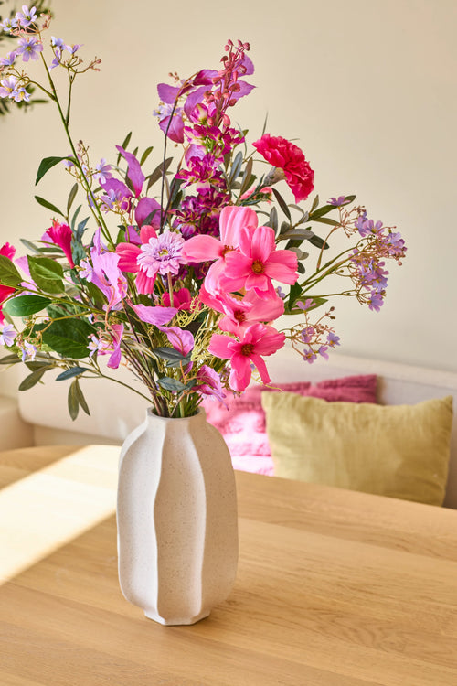 I en hvid keramikvase er 'Purple Dawn'-buketten af ​​kunstige silkeblomster, et livligt arrangement af pink, lilla og magenta blomster; vasen står på et lyst træbord med farverige puder i baggrunden.