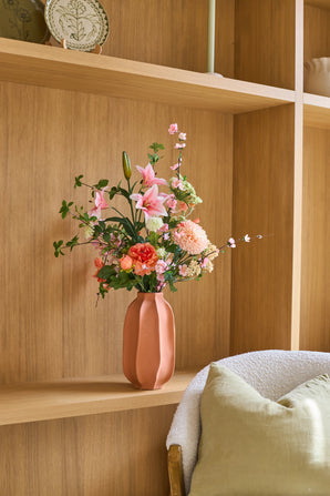 Op een houten plank naast een gezellige stoel met een groen kussen staat een roze vaas met het boeket ‘Yasmine’s Choice’ van Zijden kunstbloemen, wat een warm en uitnodigend interieurbeeld oplevert.