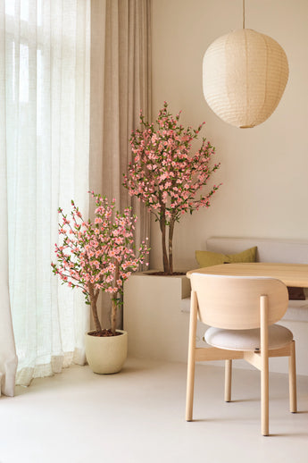 Et minimalistisk værelse med lyse træmøbler, cremefarvede gardiner og to kunstige kirsebærblomster på 150 cm i pink, som giver pink blomster. En rund papirlanterne hænger over spisebordet, med en polstret stol og indbygget bænk.