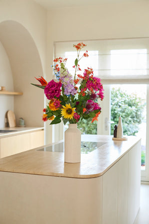 Een witte vaas met het boeket ‘Colourful Spirit’ van Zijden kunstbloemen staat op een licht houten keukeneiland in een lichte, moderne keuken met grote ramen en een neutrale inrichting, en vormt zo een duurzaam en levendig blikpunt.
