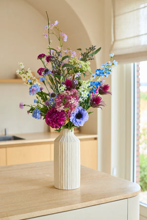 In een hoge, crèmekleurige keramische vaas staat het Zijden kunstbloemenboeket Purple Rain, een arrangement van paarse, blauwe, roze en witte zijden bloemen. Dit handgemaakte boeket staat sierlijk op een licht houten aanrecht, badend in zacht natuurlijk licht – een ware weerspiegeling van Nederlands vakmanschap.
