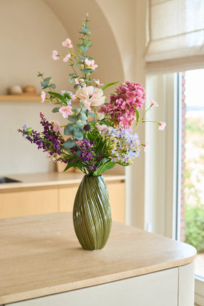 Op een lichtgekleurd keukeneiland bij een raam waar natuurlijk licht naar binnen stroomt, staat een groene vaas met roze, paarse en witte bloemen uit het Zijden kunstbloemenboeket ‘Lavender Dreams’.
