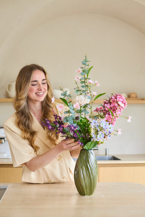 En kvinde med langt, bølget hår arrangerer en buket 'Lavender Dreams' af kunstige silkeblomster i en grøn vase i sit moderne køkken, mens hun arbejder. Den livlige buket af silkeblomster bringer tidløs skønhed til hendes hjem.