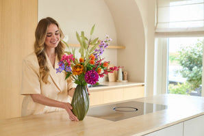 En smilende kvinde arrangerer buketten 'Artificial Silk Flowers Colorful Svenski' i en grøn vase på en lys bordplade i et køkken med moderne indretning og naturligt lys.