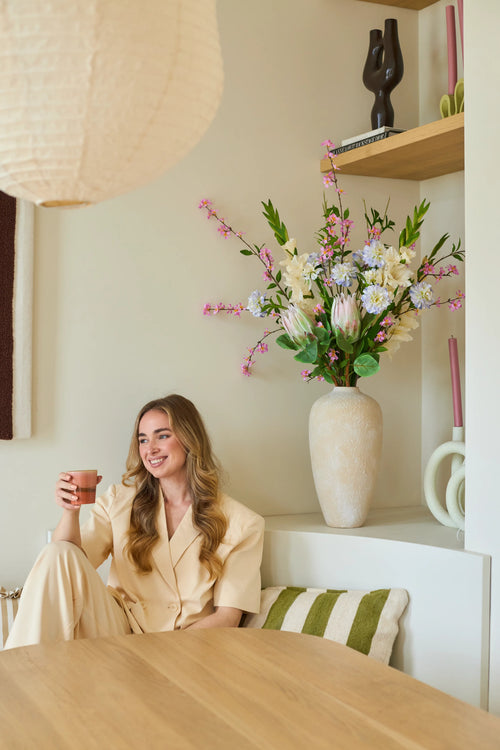 En kvinde i et cremefarvet jakkesæt sidder på en bænk med en kop i hånden og et smil på læben. Bag hende står en stor vase med den kunstige silkeblomstbuket 'Lots of Love XL', bestående af lyserøde, hvide og gule blomster, med hylder i baggrunden, hvorpå der er udstillet dekorative genstande. Rammerne ser hyggelige og moderne ud.
