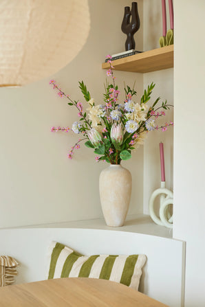 Op een witte plank staat een beige vaas met het kleurrijke boeket kunstbloemen van zijde, ‘Lots of Love XL’, naast roze kaarsen en decoratieve voorwerpen. Op een stoel onder de plank ligt een groen-wit gestreept kussen.