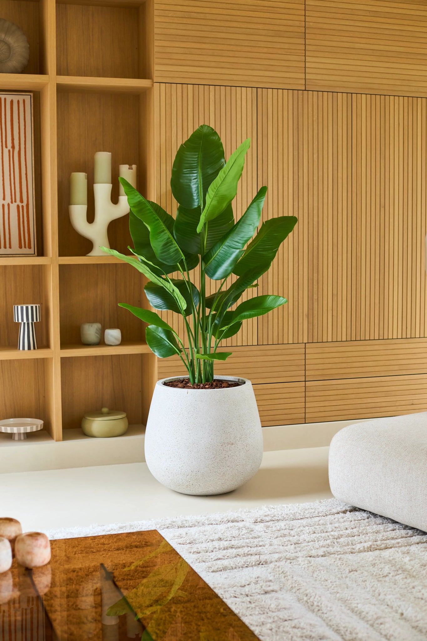Een grote Kunstplant Strelitzia Real Touch Deluxe van 150 cm in een witte plantenbak staat op een lichte vloer in een moderne woonkamer met houten lambrisering, planken en een in neutrale tinten gehouden inrichting, en geeft deze verfijnde ruimte een elegante, levensechte uitstraling.
