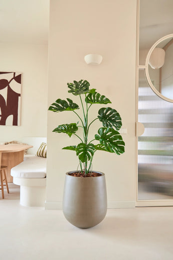 De Kunstplant Monstera van 120 cm staat in een moderne, minimalistische woonruimte met neutrale muren, een ronde spiegel, houten meubels en abstracte wandkunst.