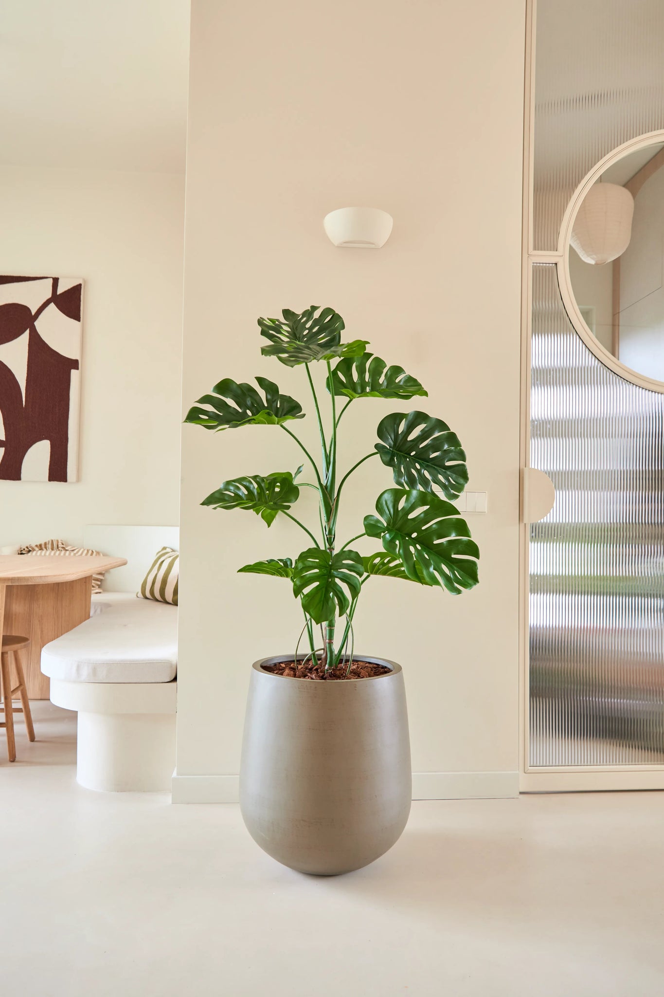 De Kunstplant Monstera van 120 cm staat in een moderne, minimalistische woonruimte met neutrale muren, een ronde spiegel, houten meubels en abstracte wandkunst.